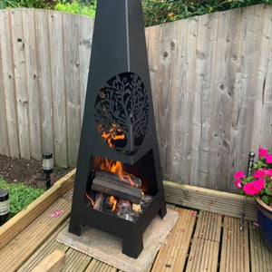Black outdoor fire pit with flames on a wooden deck, surrounded by a wooden fence and plants.